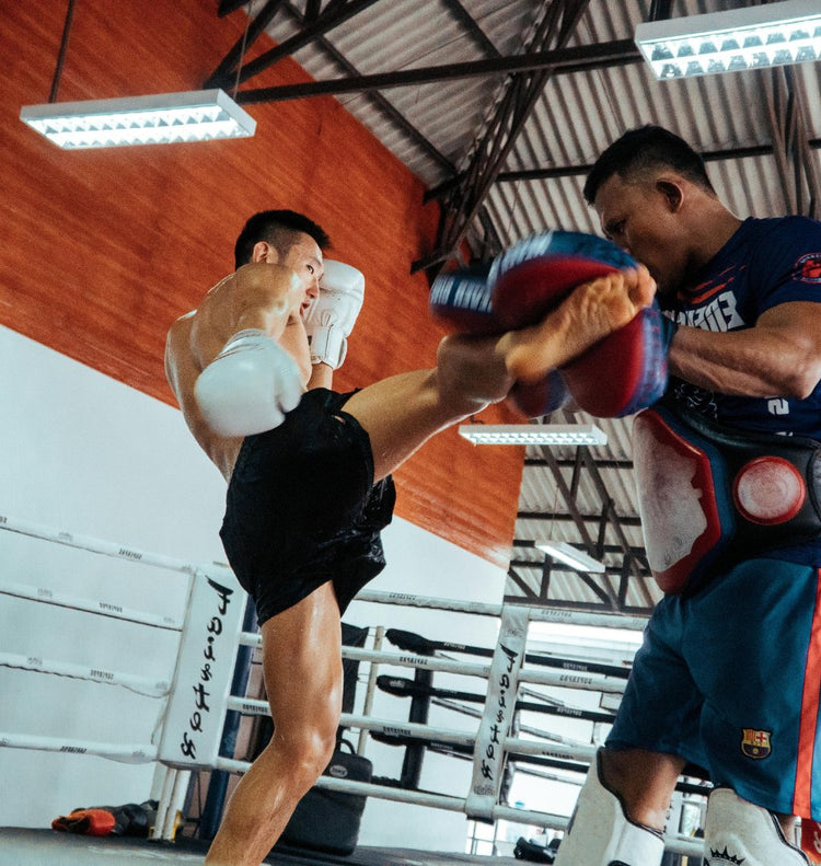Muay Thai fighter delivering a high kicking Thai Pads during training session inside a boxing gym, wearing Fairtex 16oz boxing gloves and MMA boxing shorts.