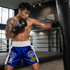 A shirtless male boxer in a gym wearing blue and white Everlast trunks and black Everlast gloves, punching a black hanging bag. The background shows a boxing ring and a wall with 'LEGENDS BOXING' written on it.