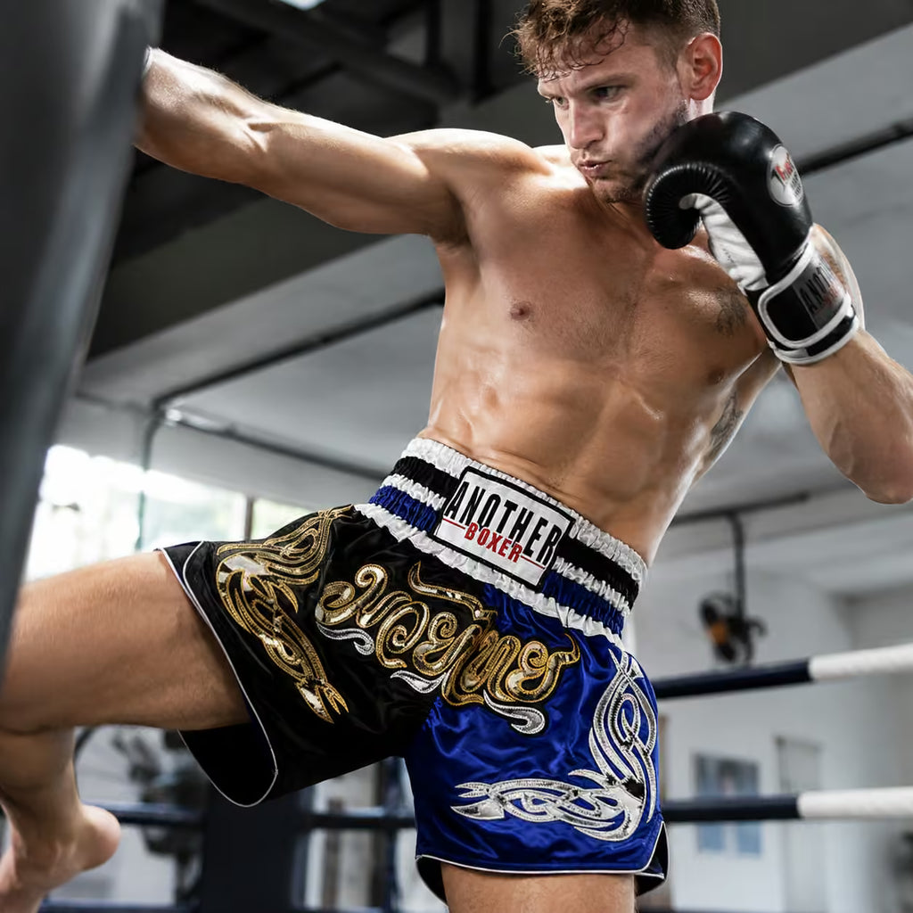 A shirtless male boxer in a ring wearing blue, black, and gold Muay Thai shorts with a white waistband reading 'ANOTHER BOXER', plus black boxing gloves, captured mid-punch in an indoor gym.
