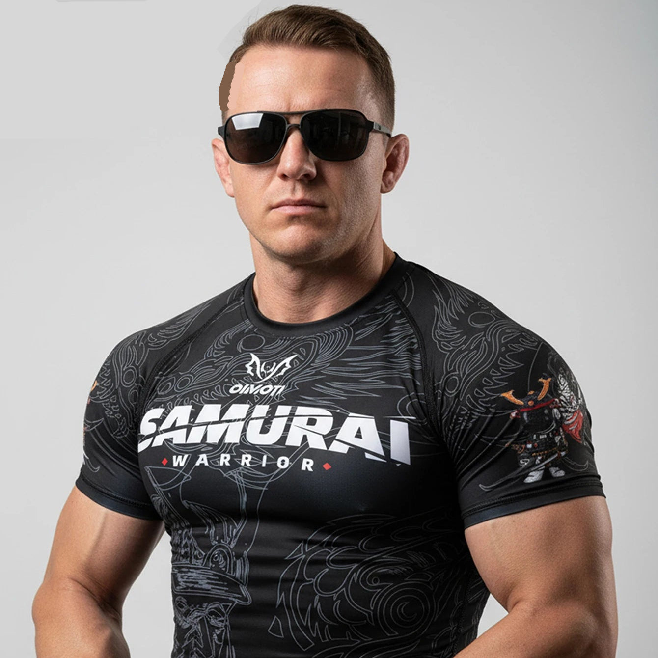 A muscular man wearing a black short-sleeve athletic rash guard with 'SAMURAI WARRIOR' printed on the chest, styled with black sunglasses. The OIMOTI brand logo is visible in the top left corner against a solid gray background.