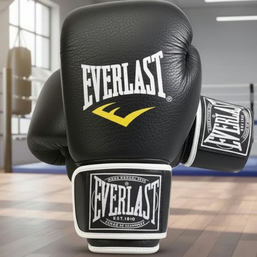 Black Everlast boxing gloves with white and yellow branding, displayed on a wooden floor inside a boxing gym.