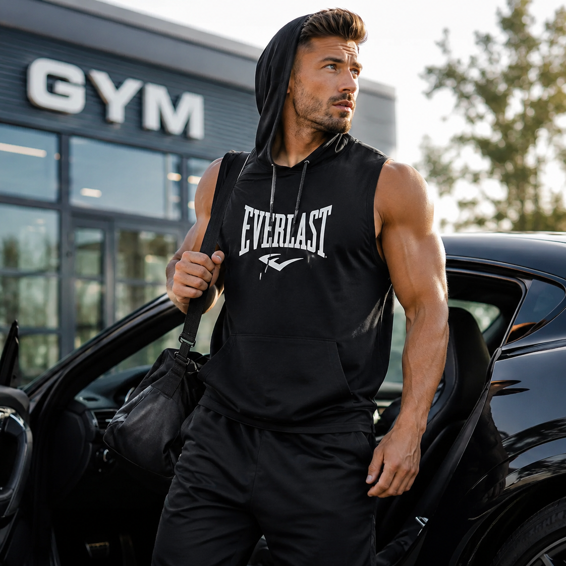 A muscular man wearing a black Everlast sleeveless hoodie and black sweatpants stands next to an open black car door outside a gym, carrying a black shoulder bag.