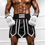 A muscular male model wearing black and white boxing shorts with white boxing gloves, standing inside a boxing ring. The brand 'ANOTHER LUXER' is clearly visible on the waistband and gloves.