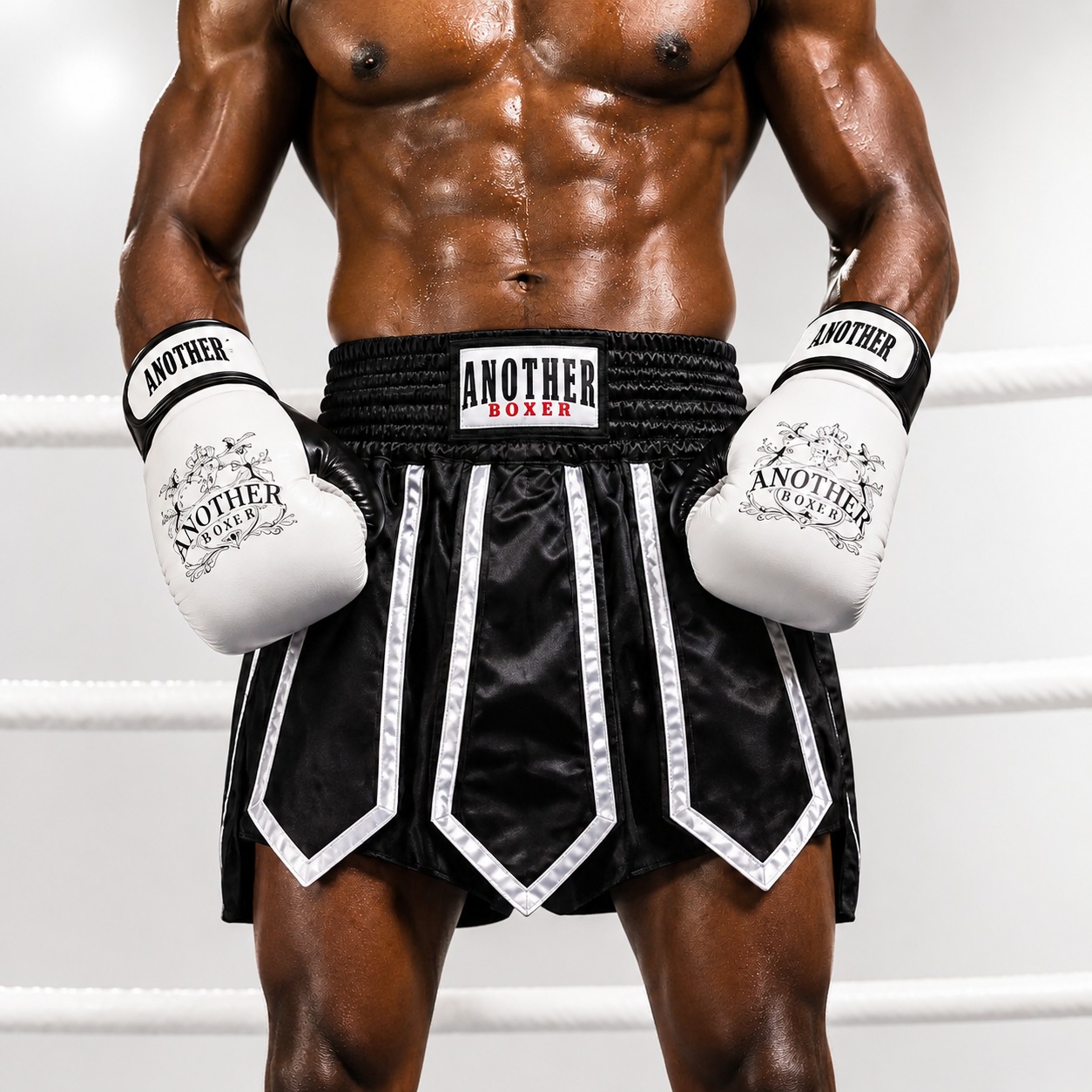 A muscular male model wearing black and white boxing shorts with white boxing gloves, standing inside a boxing ring. The brand 'ANOTHER LUXER' is clearly visible on the waistband and gloves.