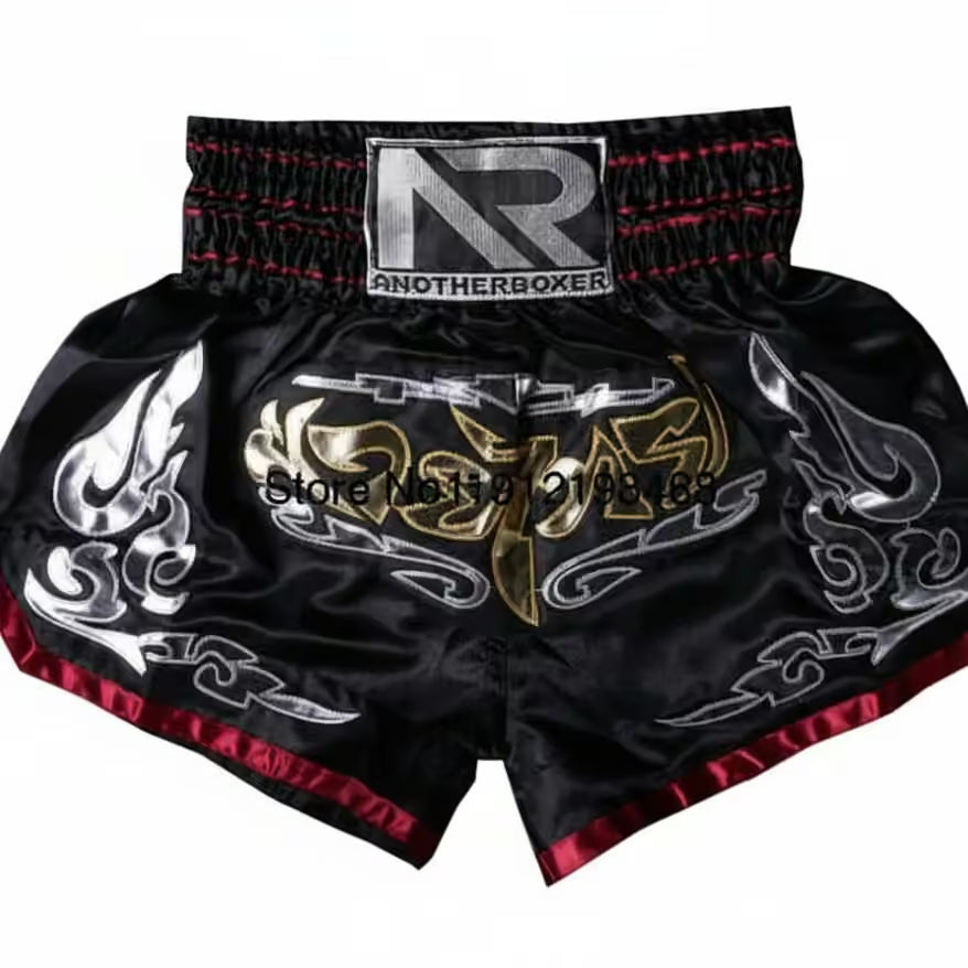 Black satin Muay Thai shorts with red trim, featuring a large gold and silver graphic on the front, a silver 'ANOTHERBOXER' brand patch on the waistband, and silver flame-like patterns on the sides.