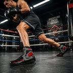 A boxer in a ring wearing black and gray boxing boots with red accents, captured in a dynamic lunge pose inside a boxing gym.