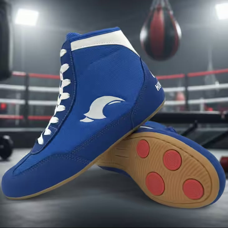 A pair of blue high-top wrestling shoes with white laces and a white logo, shown in a boxing ring with a hanging punching bag in the background.