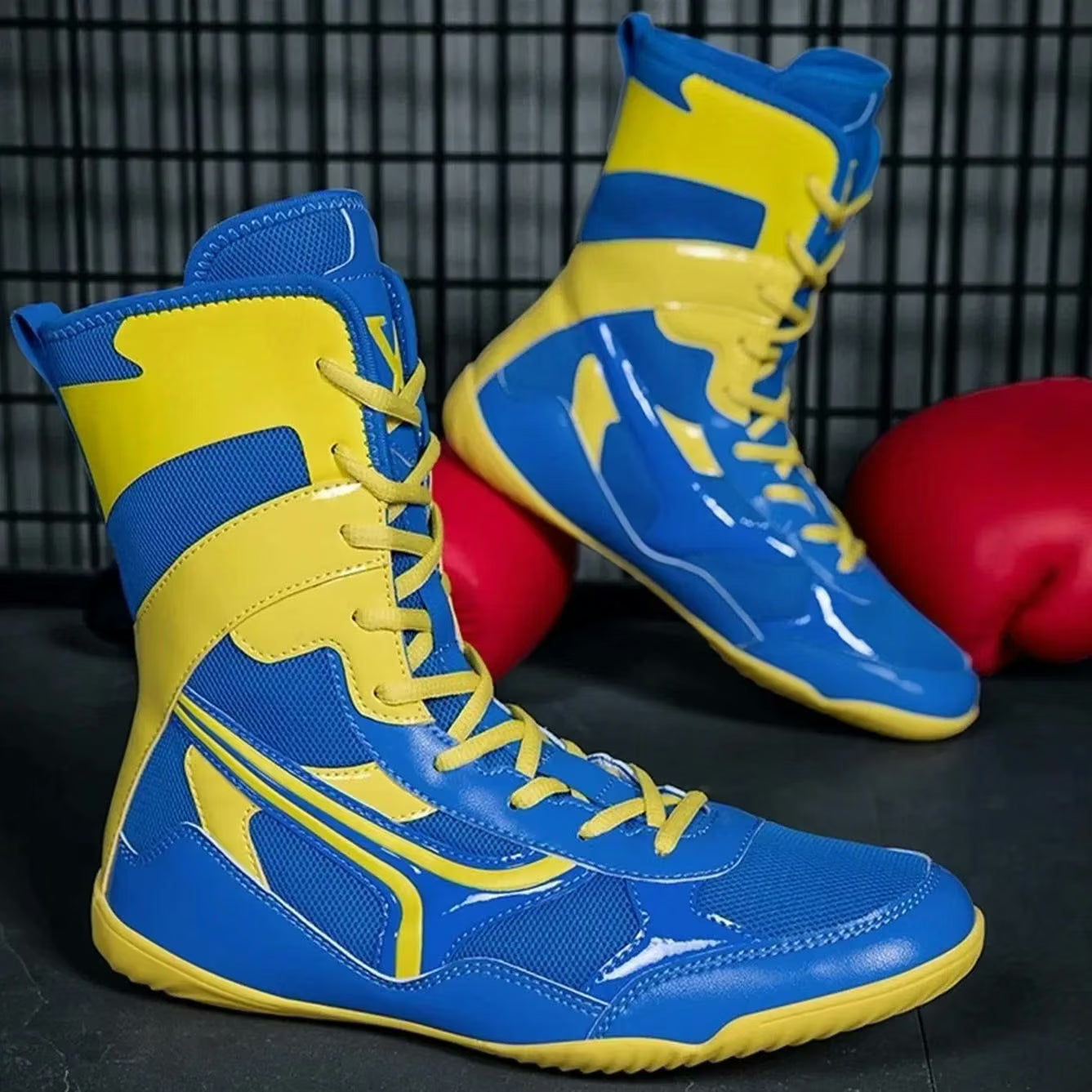 A pair of high-top boxing shoes in bright blue and yellow, displayed on a dark surface with red boxing gloves and a black wire grid background.