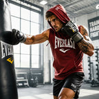 A man in a red Everlast hoodie and black shorts punches a black Everlast hanging punching bag in a gym.