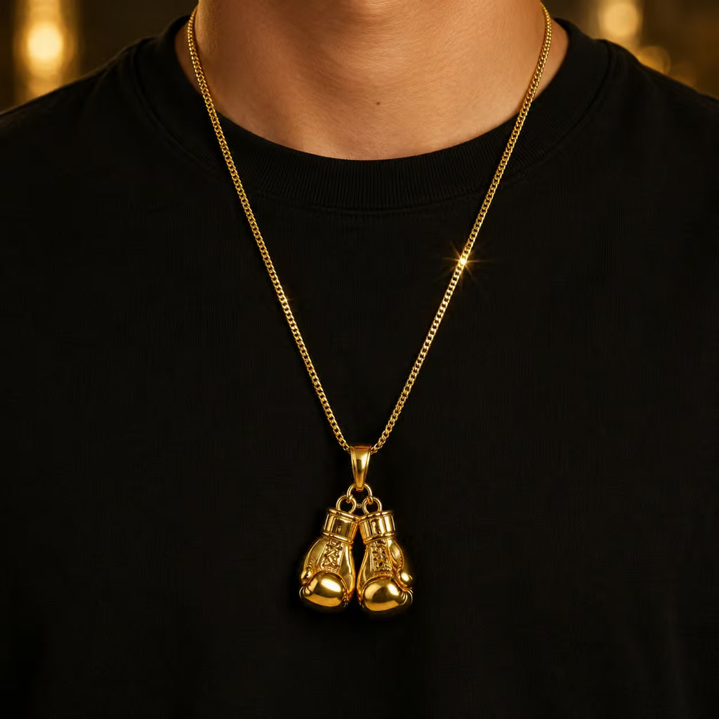 A gold pendant necklace featuring a pair of boxing gloves hanging from a gold chain, worn by a person in a black t-shirt against a warm, blurred background.