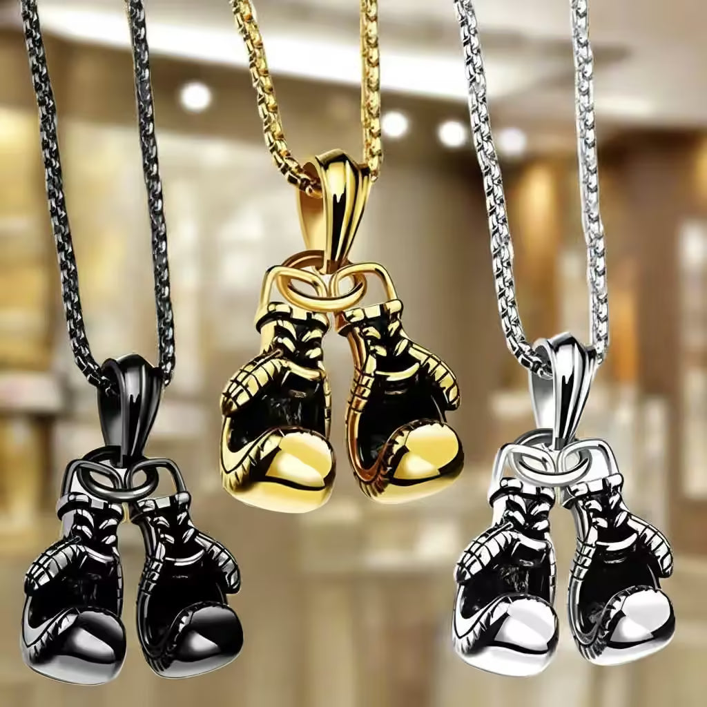 Three pairs of metallic boxing gloves pendants on chains, shown in black, gold, and silver finishes against a blurred warm indoor background.