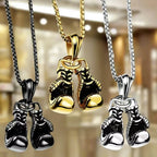 Three pairs of metallic boxing gloves pendants on chains, shown in black, gold, and silver finishes against a blurred warm indoor background.