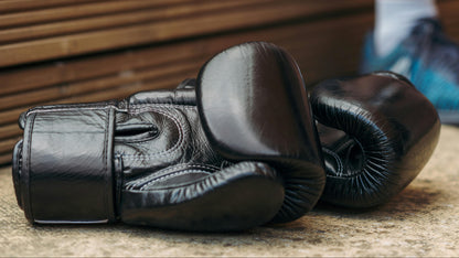 Twins Special Black boxing gloves on a concrete surface with a person wearing blue shoes in the background.