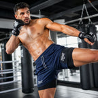 A shirtless man in a gym wearing navy and black Everlast athletic shorts and black gloves, mid-kick in a ring with punching bags in the background.
