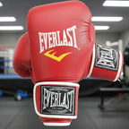 A pair of red Everlast boxing gloves with white and yellow branding, shown in a gym setting with a boxing ring blurred in the background.
