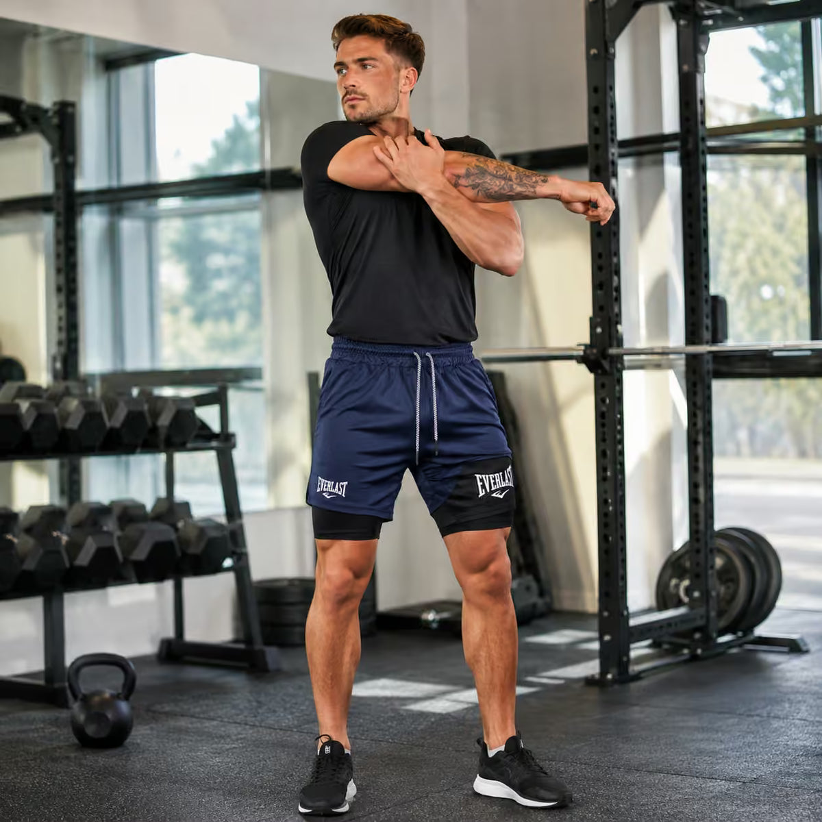 A man in a gym stretching, wearing navy blue Everlast athletic shorts and a black t-shirt. The background shows weight racks, dumbbells, and windows.