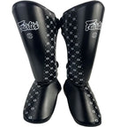 A pair of black Fairtex shin guards with white geometric patterns and white Fairtex branding, shown against a plain white background.