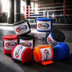 A group of Fairter hand wraps in multiple colors including blue, red, orange, black, and white, arranged on a dark gym floor with a blurred boxing ring in the background.