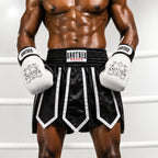 A muscular male model wearing black and white boxing shorts with white boxing gloves, standing inside a boxing ring. The brand 'ANOTHER LUXER' is clearly visible on the waistband and gloves.