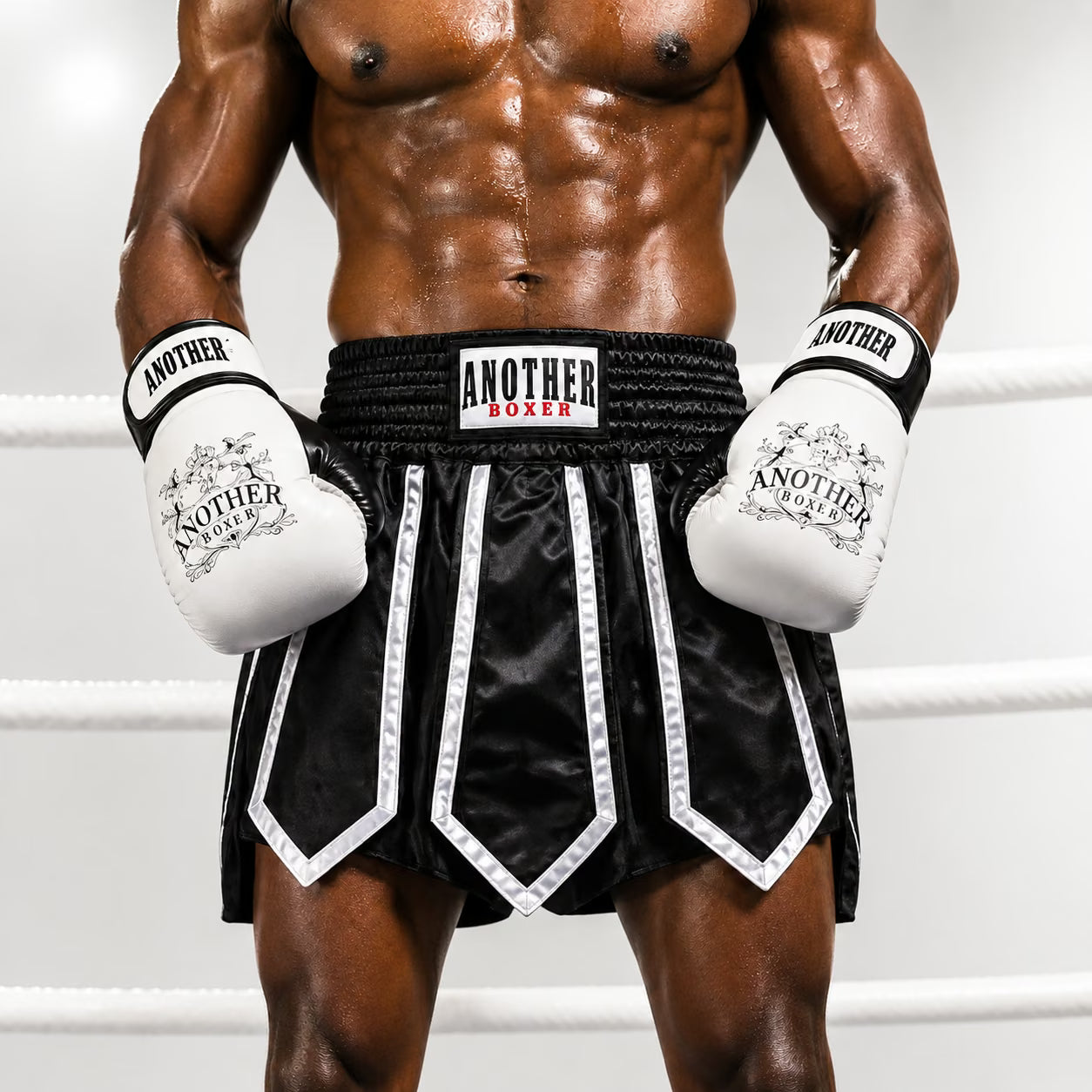 A muscular male model wearing black and white boxing shorts with white boxing gloves, standing inside a boxing ring. The brand 'ANOTHER LUXER' is clearly visible on the waistband and gloves.