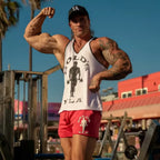A muscular man posing outdoors in a white tank top with a black graphic and red shorts, flexing his biceps. The background shows a sunny outdoor gym setting with red buildings and blue sky.