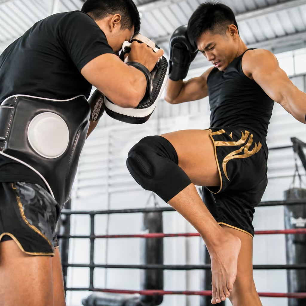 Two male athletes in a boxing ring, one wearing a black t-shirt and black shorts with gold trim while the other wears a black tank top and black shorts. The athlete on the right delivers a high kick to the athlete on the left, who is wearing a black protective headguard and wearing black gloves.