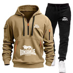 A two-piece Lonsdale tracksuit set featuring a tan pullover hoodie with a half-zip front, kangaroo pocket, and black drawstrings, paired with black jogger pants with white Lonsdale branding and white sneakers.