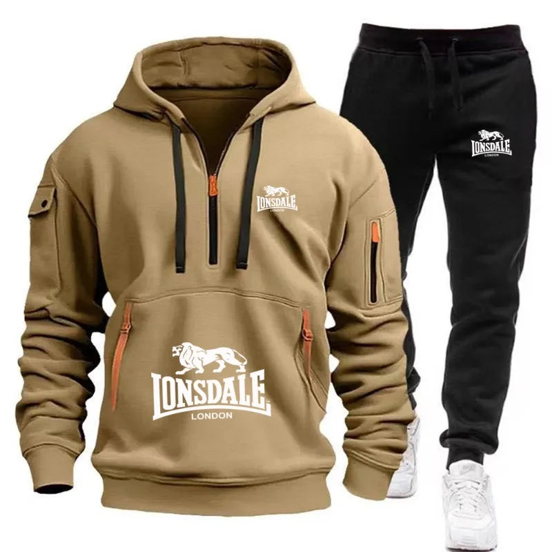 A two-piece Lonsdale tracksuit set featuring a tan pullover hoodie with a half-zip front, kangaroo pocket, and black drawstrings, paired with black jogger pants with white Lonsdale branding and white sneakers.
