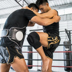 Two martial artists grappling inside a boxing ring, wearing black athletic gear including shorts with gold trim, a black belt, and a knee pad, with punching bags visible in the background.