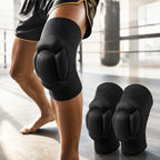 Black knee pads with ribbed protective panels worn on a person's knee in a boxing ring, with a second pair placed on the floor nearby.
