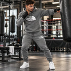A man in a boxing gym wearing a heather gray The North Face hoodie and matching sweatpants, punching a heavy bag. The hoodie features a white 'THE NORTH FACE' logo on the chest, and the sweatpants have a small white logo on the thigh.