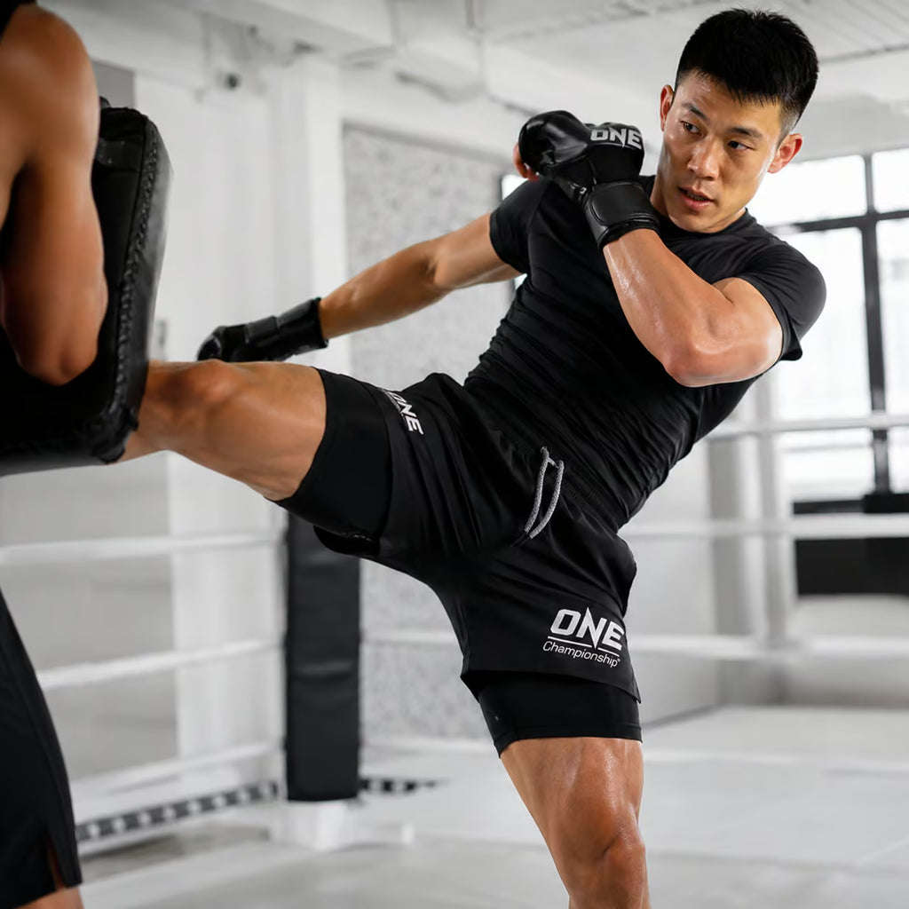 A male athlete in a boxing ring wearing a black ONE Championship athletic kit, including a fitted t-shirt, shorts with the ONE logo, and boxing gloves, while delivering a high kick toward a black pad held by another person.