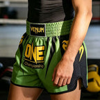 Green and black Venum Muay Thai shorts with yellow 'ONE Championship' branding, worn by a model in a gym setting.