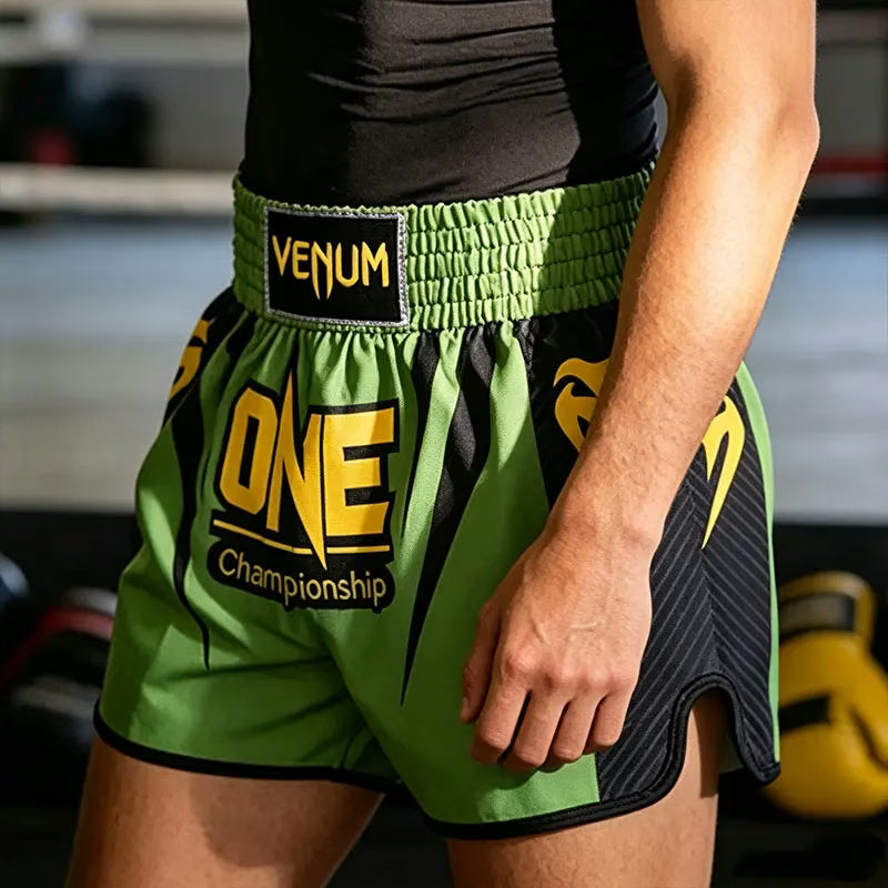 Green and black Venum Muay Thai shorts with yellow 'ONE Championship' branding, worn by a model in a gym setting.