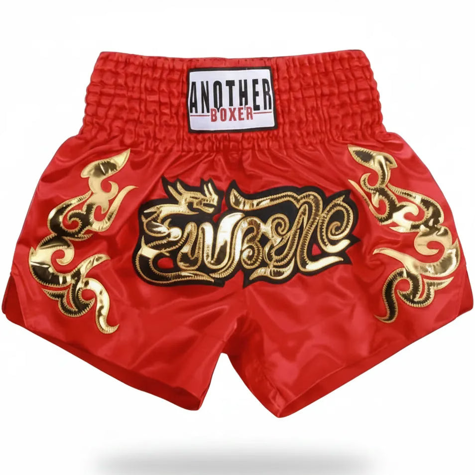 Red Muay Thai boxing shorts with gold and black embroidered graphics, featuring a white waistband label that reads 'ANOTHER BOXER'.