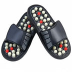 A pair of black slide sandals with massage nodes on the soles. The soles feature rows of small white and red spherical massage beads set into a black textured footbed, with a wide black strap across the top.