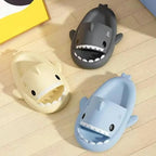 Three shark-shaped slippers in gray, beige, and blue on a wooden floor.