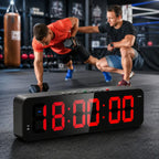 A black digital workout timer with large red LED numbers sits in the foreground on a gym floor, with two men exercising in a boxing gym behind it.