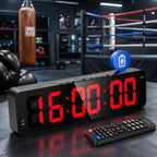 A black digital alarm clock with large red LED numbers showing 16:00:00, placed in a boxing gym setting. The clock has a remote control, a blue battery icon overlay, and a punching bag and boxing ring in the blurred background.