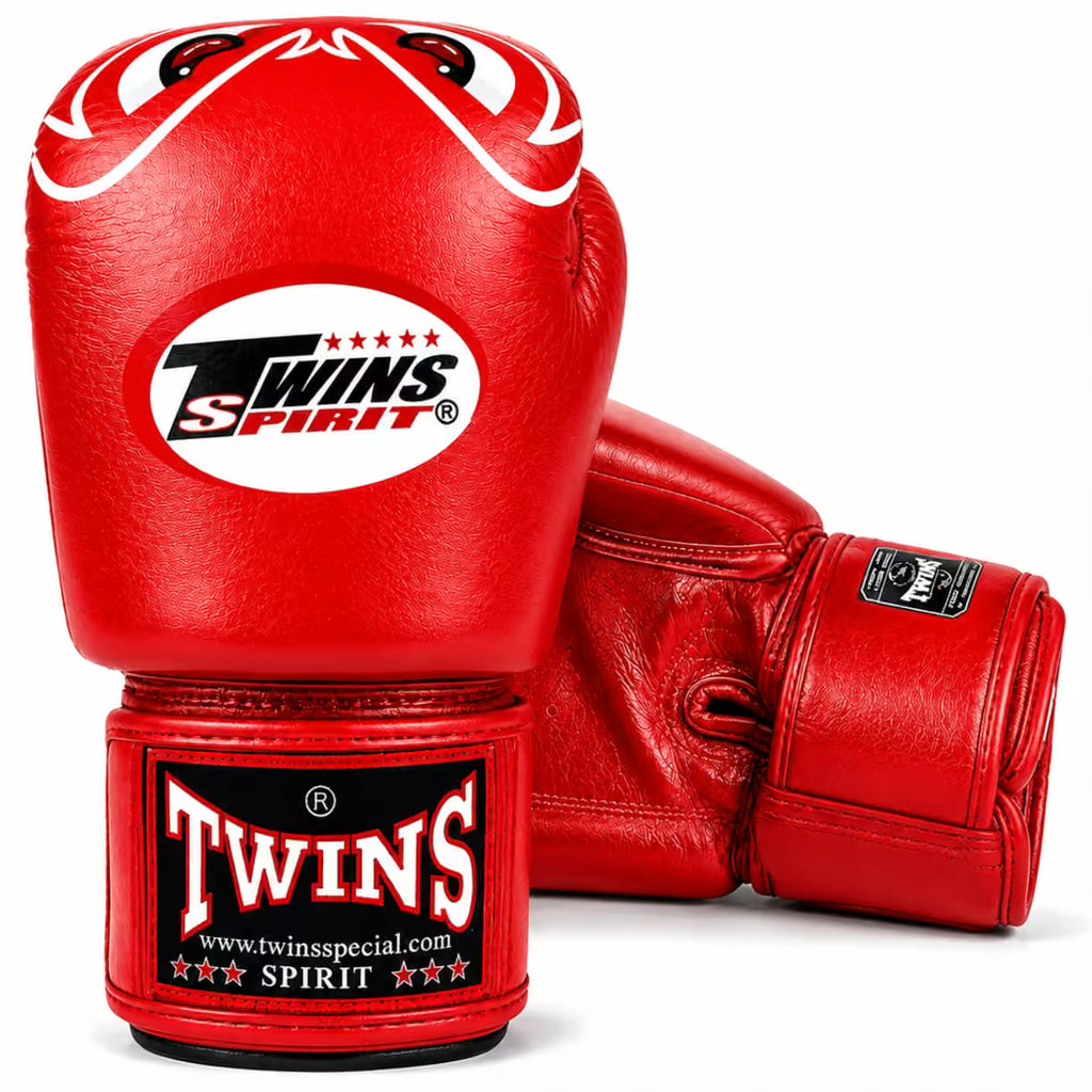 A pair of red leather boxing gloves with white and black branding. One glove is upright, showing the Twins Spirit logo on the knuckle area and a Twins brand wrap around the wrist.