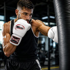 A male boxer in a black tank top and shorts throws a punch against a black heavy bag in a gym, wearing Twins brand white boxing gloves.