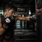 A male boxer in a black tank top and Twins boxing gloves delivers a punch to a black Twins heavy bag in a dimly lit boxing ring.