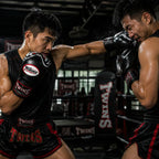 Two male boxers in a training ring wearing Twins boxing gear, with one boxer delivering a punch to the other in a dynamic action shot.