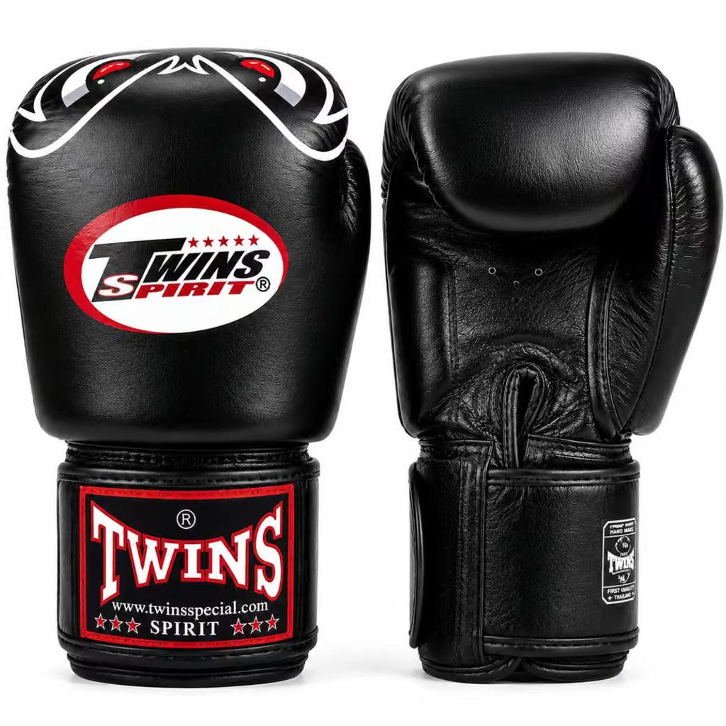 A pair of black leather boxing gloves shown from the front and back on a white background. The front glove features a red, white, and black 'Twins Spirit' logo patch on the knuckle and a matching red and white 'Twins' brand patch on the wrist strap.