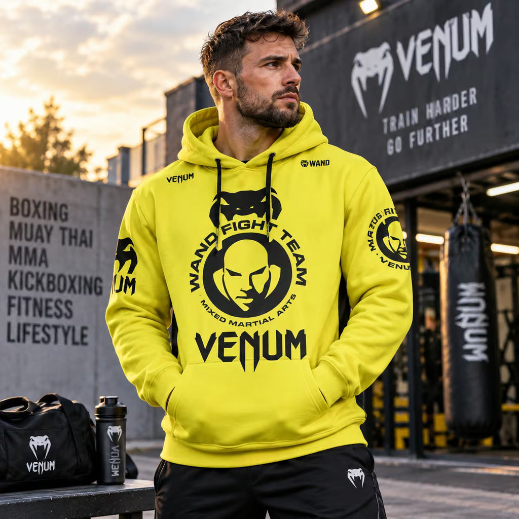 A man wearing a bright yellow Venum Fight Team hoodie with a large black graphic on the front, standing in a Venum gym with punching bags and a black duffel bag on a bench.