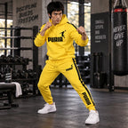 A man in a bright yellow PUMA tracksuit with black stripes and logos stands in a gym, posing in a fighting stance. The tracksuit includes a pullover hoodie and matching jogger pants, paired with white Puma sneakers.